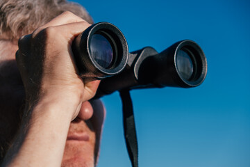 man stands outdoors, using binoculars to carefully observe surroundings. bright blue sky provides perfect backdrop for this serene activity in nature. close up.