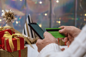 person enjoys online shopping during holiday season. they hold credit card in one hand while looking at phone, surrounded by beautifully wrapped gifts and festive decorations. close up.