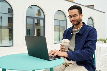 Young Black man sitting outdoors using laptop and holding disposable coffee cup, working or studying at round table in modern courtyard, looking at screen with slight smile