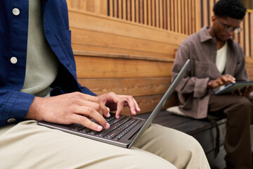 Closeup of young man typing on laptop while Black young man using digital tablet in background, both sitting on wooden bench in modern setting