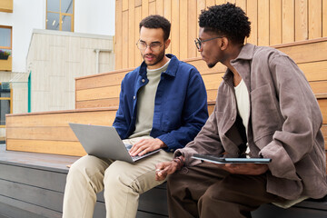 Two young men sitting on outdoor wooden steps, collaborating while using laptop and digital tablet, discussing project or studying together