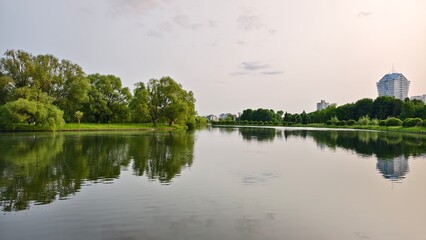 In the evening, the summer sun sets over the horizon, illuminating the buildings, grassy shores of the lake, willows, and other trees. The water ripples and reflects. The weather is clear