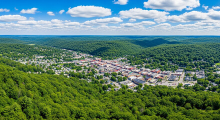 Obraz premium Aerial view of a small town nestled in a valley surrounded by lush green forests on a sunny day with clouds