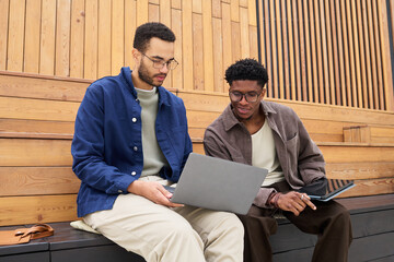 Two young Black men sitting on wooden benches using laptop and digital tablet, collaborating on project or studying together, both focused on screen, casual outdoor setting
