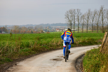 Fototapeta premium Cyclist in colorful gear rides a dirt road through green fields on an overcast day, with a distant village and bare trees in the background.