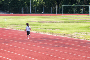 Fototapeta premium A little girl runs alone on the red tartan track of a small stadium during a sunny day