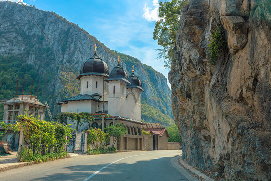 Mraconia Monastery nestles at the base of a towering cliff above the wide Danube, with boats moored along the rocky shore and dramatic gorge scenery in view in Romania