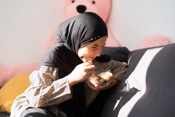 Young happy girl in hijab sitting on sofa eating a cereal meal, cozy home setting with soft sunlight and teddy bear in background.