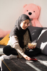 Young girl in hijab sitting on sofa, holding a bowl of cereal and using a smartphone, cozy home setting with soft sunlight and teddy bear in background.