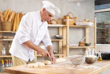 Elderly experienced male baker using knife to cut raw dough to make croissants