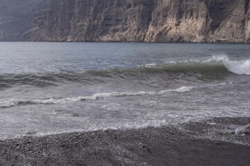 Fototapeta premium Ocean wave rolling on black volcanic sand beach with foamy surf and reflection of cloudy sky near Los Gigantes, Tenerife, Canary Islands, Spain. Beautiful seascape nature background.