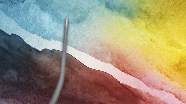 A brush skillfully applies vibrant watercolor paint to textured paper, creating a dynamic blend of deep blues, warm yellows, and rich maroon tones. The fluid strokes and organic edges suggest a creati