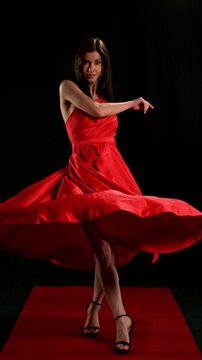 Stunning Black-Haired Woman in Ruby Red Dress and High Heels Gracefully Posing Against a Black Background, Captured in Super Slow Motion. Vertical Aspect Ratio. Speed Ramp Effect.