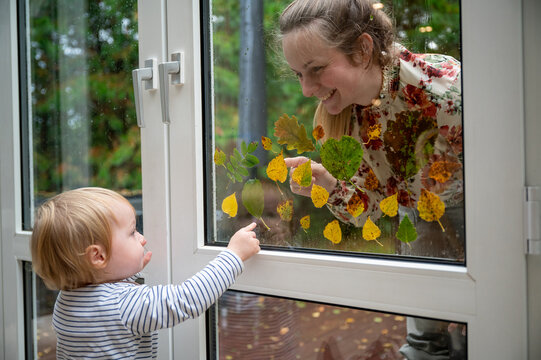Young mother and her 15-month-old son playing creatively outdoors on a rainy day, decorating the window with colorful autumn leaves, learning and bonding through nature-inspired play