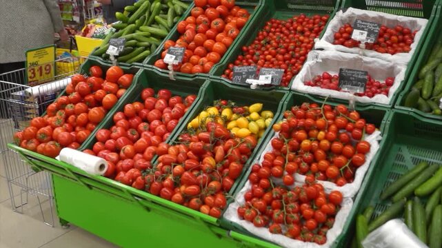 Colorful tomato varieties and fresh cucumbers on display at supermarket in Russia, with shopping cart and price tags visible.
