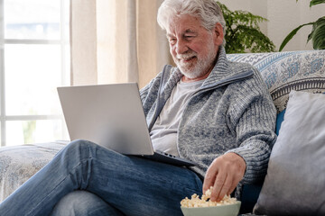Elderly 70s man seated on sofa browsing on laptop, senior looking at laptop screen surfing the net while eating popcorn, older generation and modern tech application easy usage concept