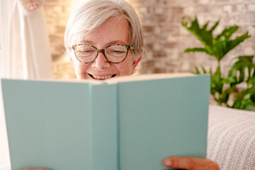 Portrait of smiling caucasian senior woman with eyeglasses partially hidden by a book enjoying pleasure of reading - concept of relaxed lady and retirement lifestyle