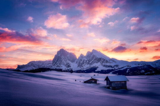 Snowy Mountain Sunset Over Alpe di Siusi With Cabins and Soft Pink Clouds A tranquil winter scene shows snow-covered fields and rustic cabins beneath towering alpine peaks as the sun sets, painting...