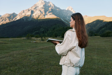 A thoughtful woman stands in a scenic mountain meadow, holding a tablet and looking toward the...