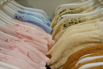 Soft pastel pink blue yellow cream baby clothes neatly arranged on white plastic hangers in a boutique store, highlighting delicate embroidery and gentle cotton texture for newborn girls.