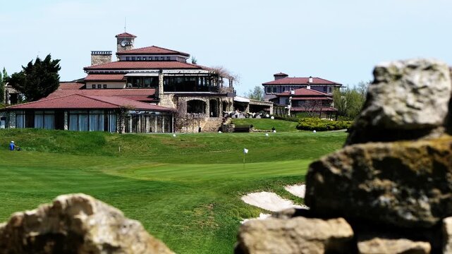 Distant static shot of golfers playing and driving carts at BlackSeaRama golf complex in Bulgaria, showing the clubhouse, clock tower, green field, and sea view.