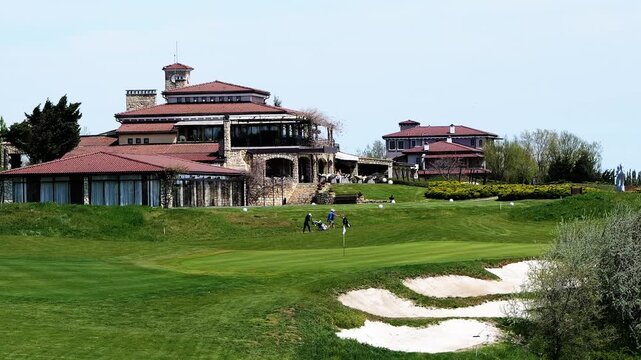 Distant static shot of golfers playing and driving carts at BlackSeaRama golf complex in Bulgaria, showing the clubhouse, clock tower, green field, and sea view.