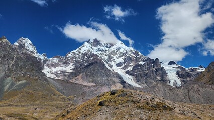A imagem mostra uma imponente montanha coberta de neve e gelo, erguendo-se sob um c&eacute;u azul profundo com nuvens dispersas. As encostas rochosas e &aacute;ridas contrastam com o brilho branco das geleiras que 