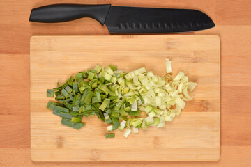 An overhead shot features a pile of chopped leeks, transitioning from dark green to white, on a bamboo cutting board with a black-handled knife above it, all on a wooden tabl