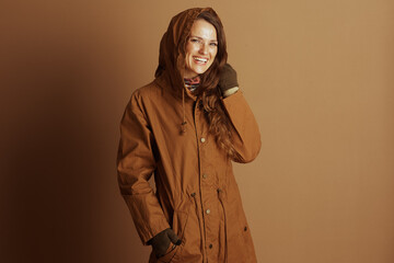 A cheerful woman with long brown hair smiles, wearing a brown hooded jacket, green gloves, and a vibrant, colorful scarf, epitomizing cozy autumn or winter fashion.