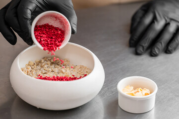 Close-up of gloved hands adding freeze-dried raspberries to oatmeal in a white bowl, with sliced almonds nearby. Food preparation scene on stainless steel surface.