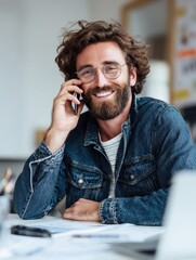 Happy digital professional in casual outfit talking on phone, using laptop on white table, working remotely from home office.