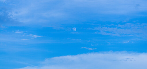 Sky and clouds with the moon visible during the day,Clear blue sky with fluffy clouds and a crescent moon visible in the background during the late afternoon hours.