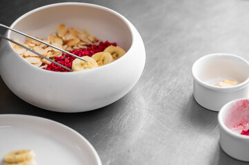 Close-up of healthy breakfast preparation with banana slices, almonds, and freeze-dried raspberries in a white bowl, arranged on a metal surface with kitchen tools and ramekins