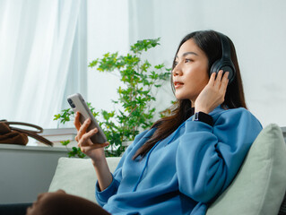 Young Asian woman Relaxing at home wearing headphones Listening to Music Streaming on Mobile Phone app sitting on sofa. Asian female using smartphone listen to podcast, browsing social media at home.