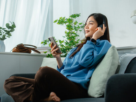 Young Adult Asian woman Listening to Music on Mobile phone Relaxing at home. Cheerful Asian woman Wearing headphones Enjoying holiday moment listening podcast, chatting on smartphone sit on sofa.
