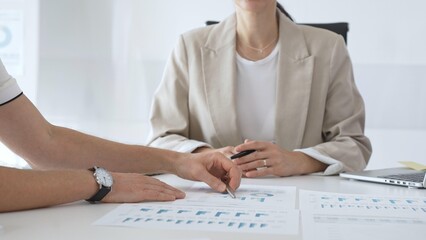 Professional businesspeople collaborating on financial analysis, female executive gesturing toward charts while presenting strategic insights to team members in corporate workspace