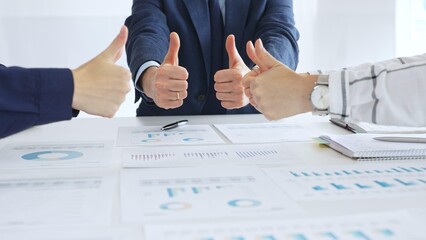 Business team showing thumbs up sign in meeting celebrating success with financial charts, data analysis, sales growth reports, and teamwork achievement in modern office
