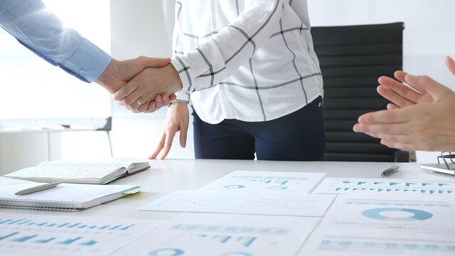 Business partnership success concept with handshake in modern office, financial charts, graphs and laptop on desk representing teamwork, agreement and corporate growth