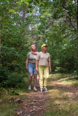 Two older women walking on a dirt path in a lush green forest, one woman placing a hand on the...