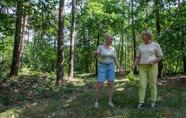 Fototapeta premium Two mature female friends are engaged in conversation and smiling while enjoying a leisurely walk in a vibrant green forest, finding joy and connection in nature during their outdoor activity