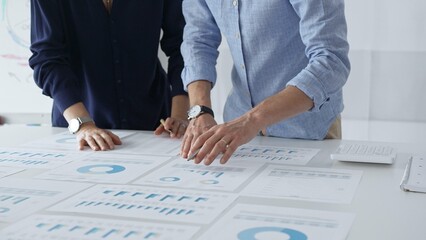 Business colleagues are examining financial data, pointing at charts and graphs spread out on a table during a collaborative analysis session in a modern office.