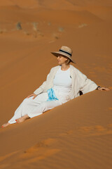 A girl is sitting on a dune in the Sahara Desert, wearing a white dress and a hat.