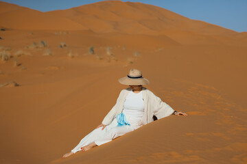 A woman wearing a hat and white clothes is sitting on the dunes of the Sahara Desert