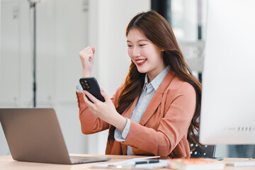 Young woman holding smartphone smiling in office setting celebrating success with laptop and desktop computer visible
