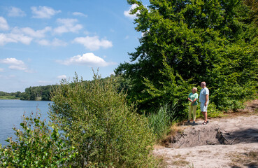 Senior couple standing by a tranquil lake surrounded by lush green trees, enjoying leisure time and watching nature during their summer vacation outdoors
