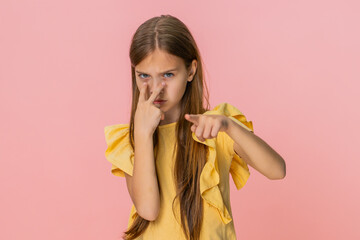 Young child girl imitates a serious expression and uses a watching you gesture, pointing two fingers from eyes toward the camera with a focused look. School kid on pink background, playful expressive.