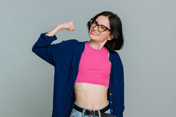 Young woman confidently raises arms, flexes biceps with proud smile, demonstrating strength and readiness to overcome challenges. Girl expresses empowered determined mood, isolated on gray background