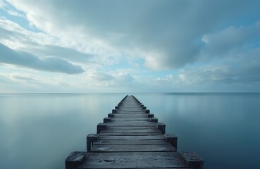 Fototapeta premium Long wooden pier extends into calm blue ocean water. Still sea reflects soft cloudy sky. Serene nature scene shows path leading to horizon. Quiet destination promotes peace, freedom, ideal for