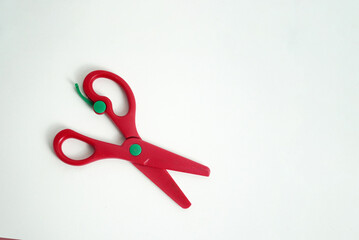 A pair of red plastic safety scissors with green pivot points, lying open on a clean white background. safe scissors for children learning to cut. school, crafts, art projects, and office supplies.