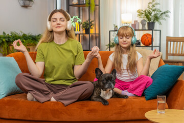 Mother and daughter with French bulldog at home use wireless headphones play calming sound start meditation. Woman and girl with dog on sofa breathe steady relax hold hands share peace warm family